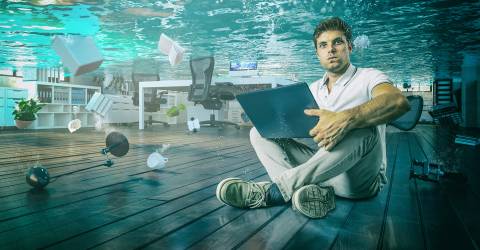 Man zit onder water gehurkt op een vloer met een laptop op schoot en  zie je een lamp, bureau, plant, ordner en boek drijven